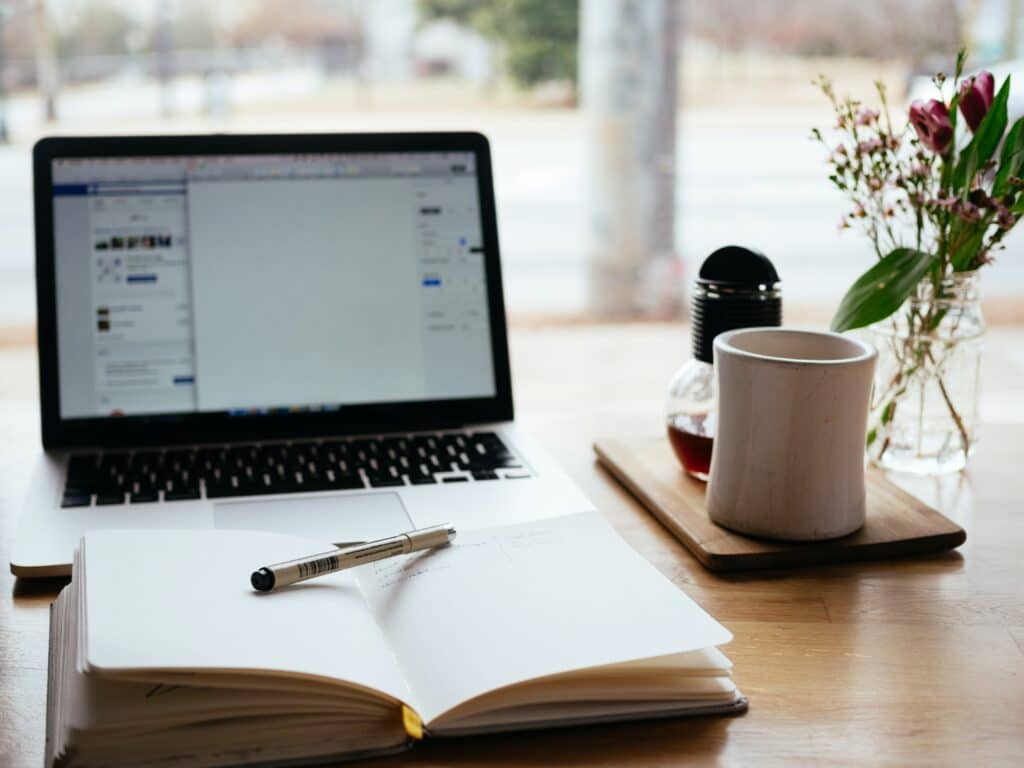 computer, journal with pen on top and coffee cup